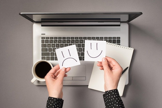 First person top view photo of hands holding two sticker notes with drawn happy and sad faces over open grey laptop pen copybook and cup of drink on isolated grey background