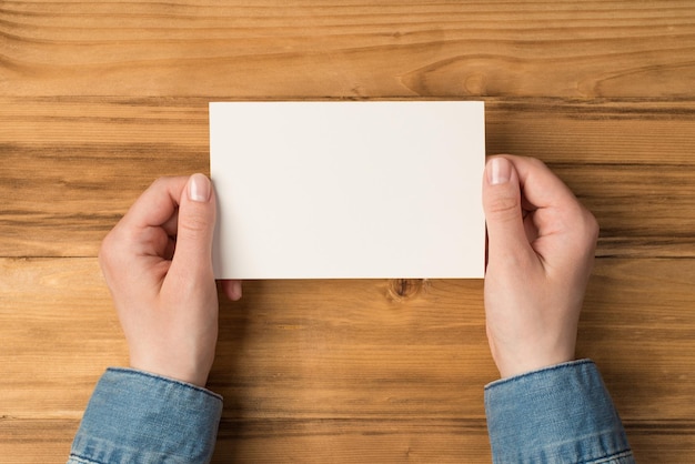 First person top view photo of hands holding white paper card on isolated wooden table background with blank space