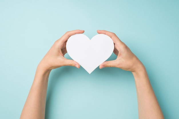 First person top view photo of hands holding white paper heart on isolated pastel blue background with copyspace