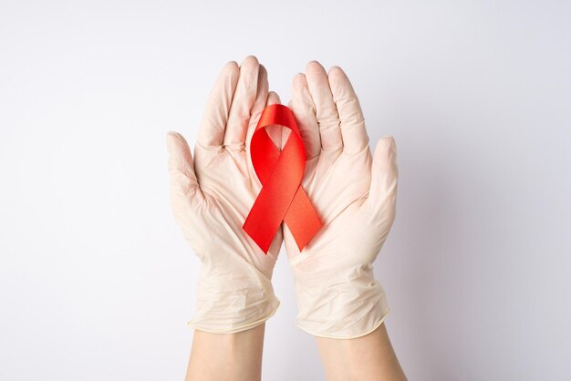 Foto in prima persona vista dall'alto delle mani in guanti protettivi medici che tengono il nastro rosso nei palmi simbolo della consapevolezza dell'aids su sfondo bianco isolato