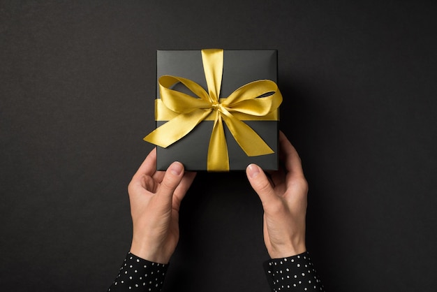 Foto in prima persona vista dall'alto delle mani in camicia a pois che danno una confezione regalo nera con fiocco di nastro di raso dorato su sfondo nero isolato