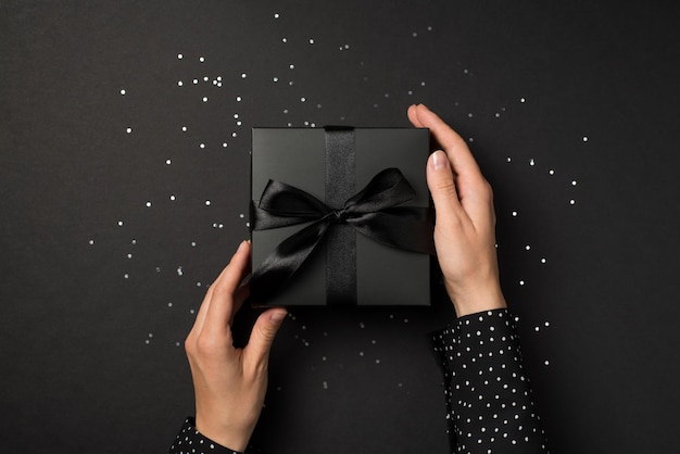 First person top view photo of hands in polka dot shirt touching stylish black giftbox with black ribbon bow over sequins on isolated black background