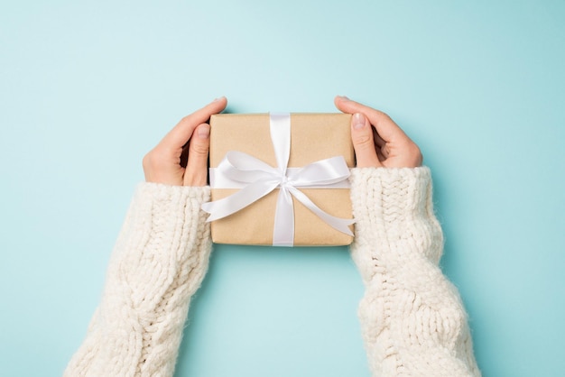 First person top view photo of hands in white knitted sweater holding craft paper giftbox with white ribbon bow on isolated pastel blue background