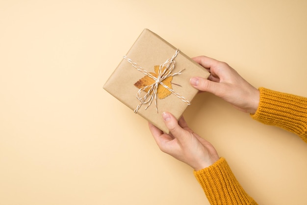 First person top view photo of hands in yellow pullover giving craft paper giftbox with twine bow and yellow autumn leaf on isolated beige background with copyspace