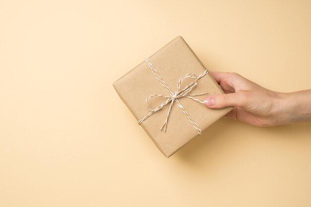 빈 공간이 있는 격리된 베이지색 배경에 꼬기 활이 있는 공예 종이 선물 상자를 들고 있는 여성의 1인칭 평면도 사진