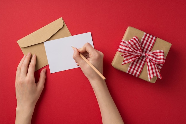 First person top view photo of st valentine's day decor female hands writing letter envelope and craft paper giftbox with checkered ribbon bow on isolated red background with copyspace