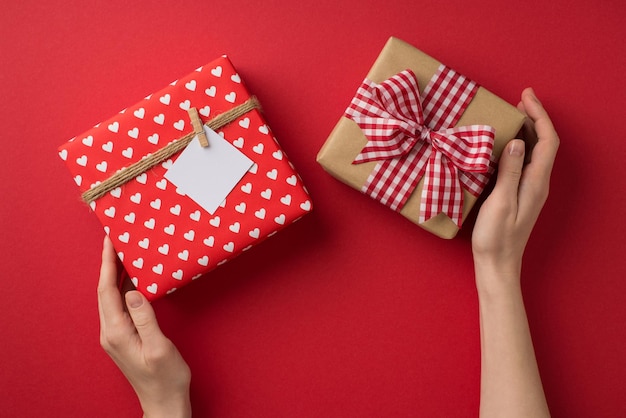 First person top view photo of valentine's day decor female hands holding craft paper giftbox with checkered ribbon bow and red giftbox with twine and pinned note on isolated red background