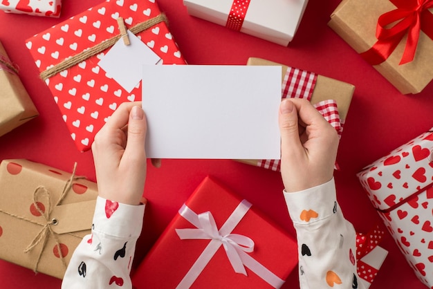 First person top view photo of valentine's day decor girl's hands in stylish white shirt holding paper sheet over present boxes on isolated red background with empty space