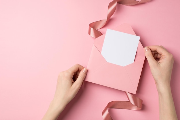 First person top view photo of valentine's day decorations female hands holding open pink envelope with paper sheet and pink silk curly ribbon on isolated pastel pink background with blank space