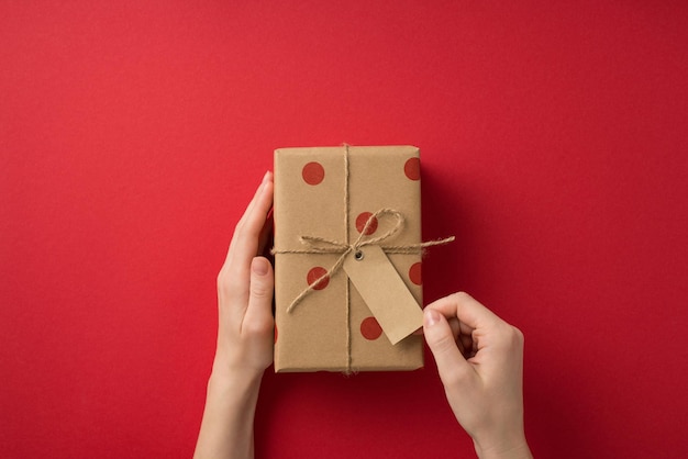 First person top view photo of valentine's day decorations woman's hands touching pricetag on craft paper giftbox with polka dot pattern and twine bow on isolated red background with empty space