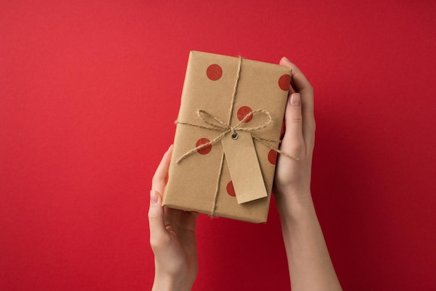 First person top view photo of valentine's day decorations young person's hands demonstrating craft paper giftbox with polka dot pattern tag and twine bow on isolated red background with copyspace