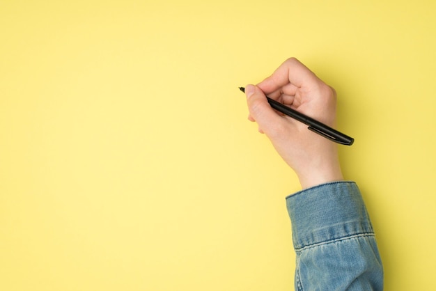 First person top view photo of woman's hand holding black pen on isolated yellow background with blank space