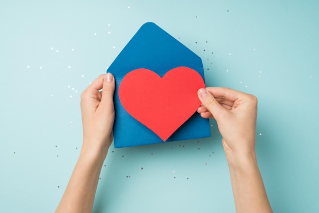 First person top view photo of woman's hands holding open blue envelope with red paper heart over shiny confetti on isolated pastel blue background with blank space