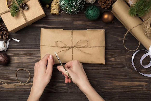 Foto foto di vista dall'alto in prima persona delle mani della donna che imballano la confezione regalo di carta kraft con le forbici dell'arco dello spago verde oro palle di albero di natale ramoscelli di pino e strumenti artigianali su sfondo di scrivania in legno isolato