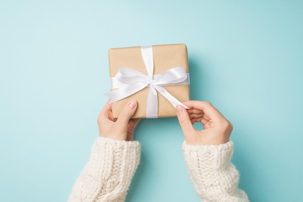 First person top view photo of woman\'s hands in white cozy\
sweater unpacking craft paper giftbox with white satin ribbon bow\
on isolated pastel blue background