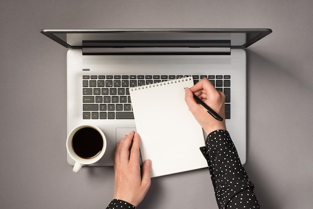 First person top view photo of woman's hands writing in notepad and cup of coffee on open laptop on isolated grey background with copyspace