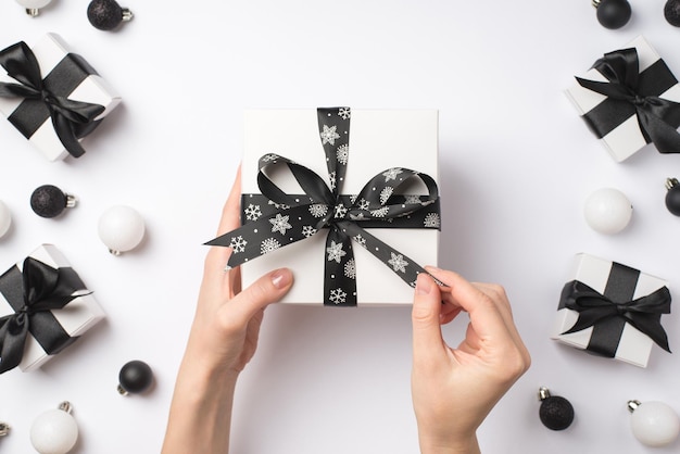 First person top view photo of young girl's hands unwrapping white giftbox with black ribbon bow over white and black christmas decorations presents and balls on isolated white background