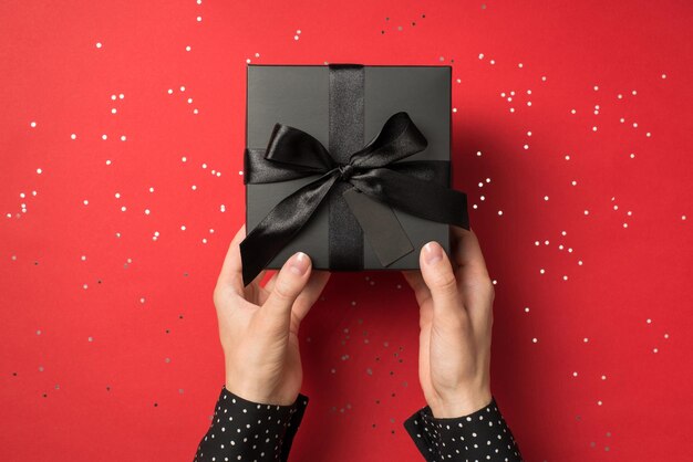 First person view above photo of black giftbox with ribbon wrapped as bow in hands and confetti around isolated on the red background