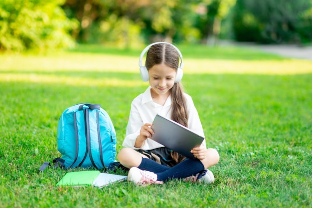 A firstgrader girl with a backpack and a tablet with headphones on a green lawn reads a book or does homework goes back to school