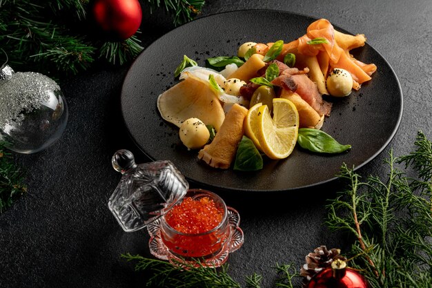 크리스마스 장난감과 가문비나무 가지가 있는 어두운 크리스마스 배경에 빨간 캐비어가 있는 검정 접시에 생선 플래터