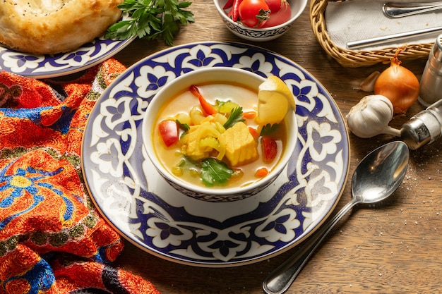 Уха из лосося, лука, помидоров, чеснока, моркови, картофеля, укропа, специй и лимона в тарелке с традиционными узбекскими орнаментами.