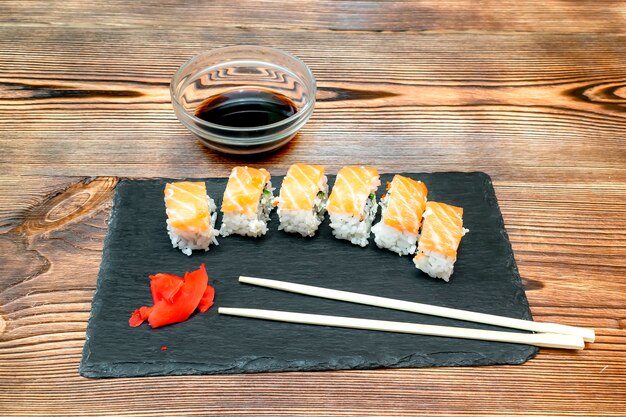 Fish sushi rolls with salmon, ginger, soy sauce and chopsticks on black cutting serving board on wooden rustic background with copy space. Seafood, Food service from restaurant concept close up.