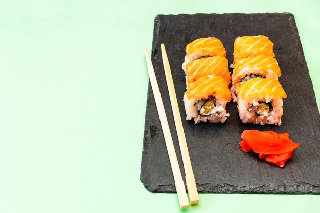Рыбные суши-роллы с лососем, васаби и палочками для еды на черной сервировочной доске. Морепродукты, Общественное питание