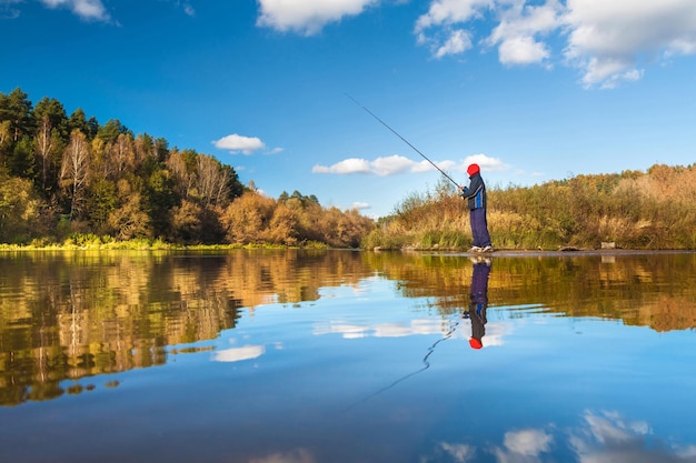 Fisherman boy on panoramic landscape with wide broad river in autumn forest in sunny day with reflection