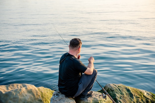 A fisherman catches fish in the sea with a fishing rod sitting on the shore of the rocks at sunset