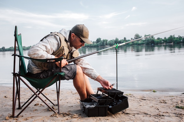 Fisherman is sitting in folding chair and leaning forward to opened black plastic box. Guy holds fly rod and plastic bait in hands. It gets chilly outside.