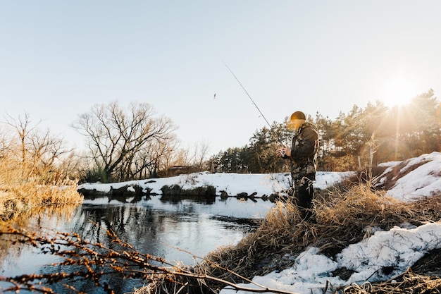 春先、雪に覆われた川のほとりで釣り竿を持った漁師が魚を釣る。