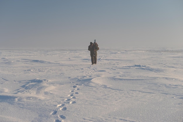 A fisherman with an ice drill and equipment walks along the surface of a frozen pond The man is talking on the phone Winter fishing theme