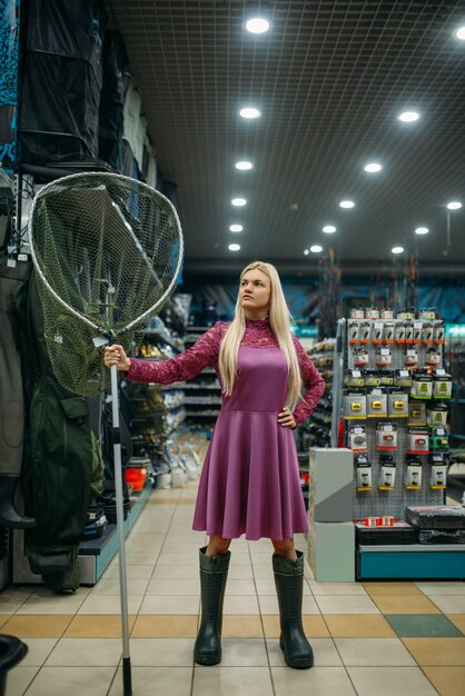 Fisherwoman in rubber boots holds net in fishing shop, hooks and baubles