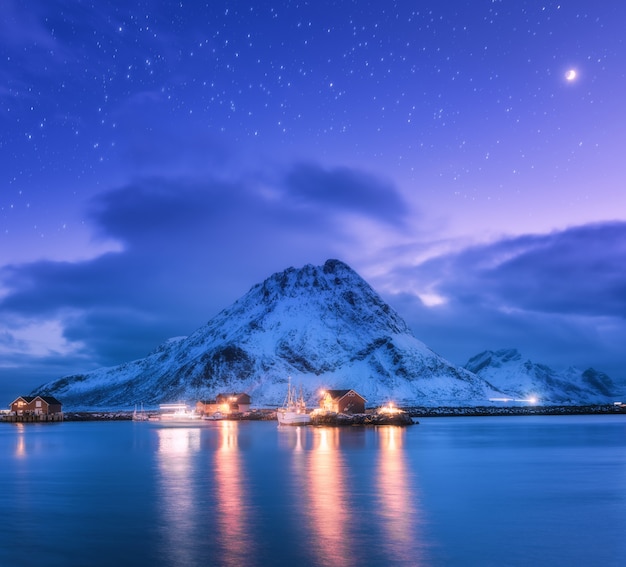 Fishing boats near pier on the sea against snowy mountains and starry purple sky with moon at night