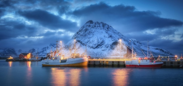 海の漁船 雪の多い岩山 ノルウェーのロフォーテン諸島の夜の雲と青い空 ボートのある冬の風景 港の街の明かり 雪の中のイルミネーションのある家