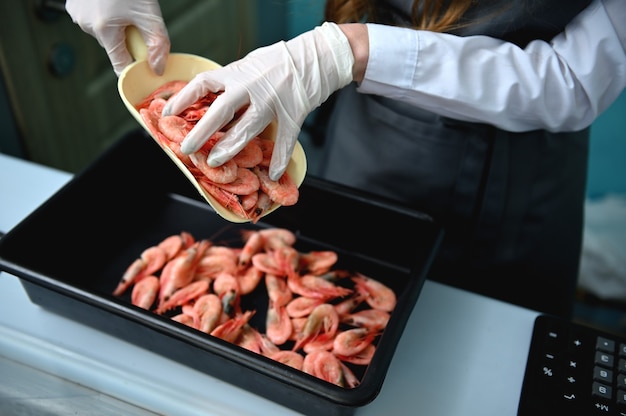 シーフードマーケットの魚部門のベンダーである魚屋は、作業用エプロンのカウンターの後ろに立ち、新鮮な冷凍の冷やした赤いエビをトレイに置きます。閉じる