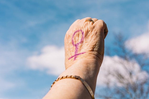 Fist of an older woman with purple painted nails with the sky in the background with the female symbol painted Concept of women39s day empowerment equality inequality activism and protest