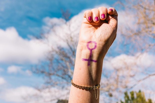 Fist of an older woman with purple painted nails with the sky\
in the background with the female symbol painted concept of\
women39s day empowerment equality inequality activism and\
protest