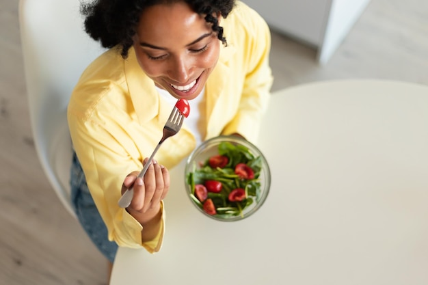 Fit african american lady eating salad with organic vegetables in light kitchen interior top view free space