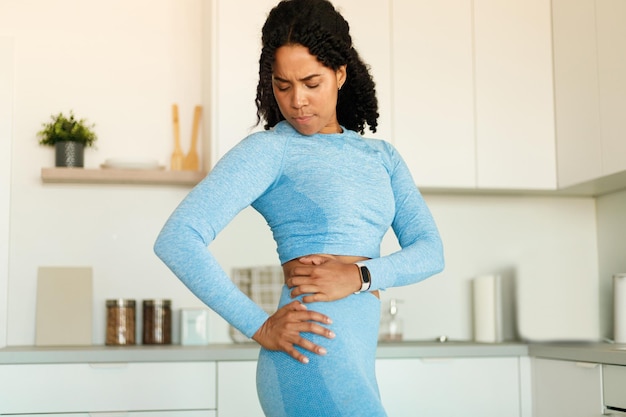 Fit african american woman having trauma back suffering from\
pain standing in kitchen interior copy space