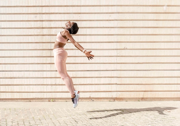 A fit african american woman jumping into the sun with her arms\
stretched back in sportswear in the street outdoor sports freedom\
spirit concept