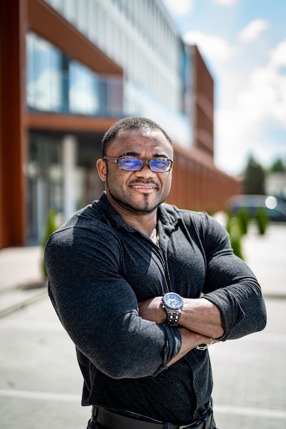 Fit African man wearing glasses and dark tight shirt Guy with big biceps and his hands folded on the massive chest Smiling strong man standing in the street in bokeh effect