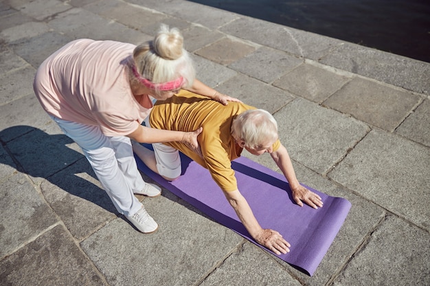 fit aged lady helping her husband performing a stretching exercise outdoors