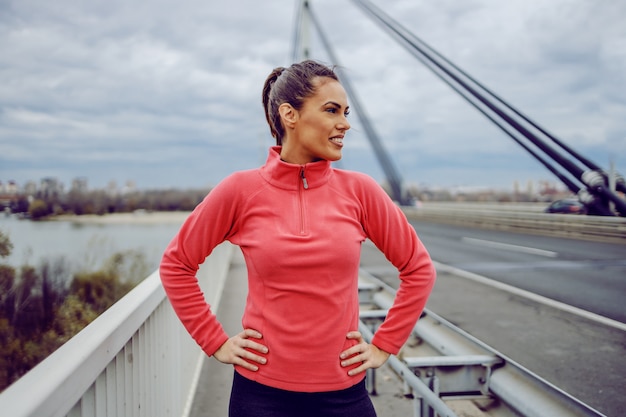 Fit attractive young sportswoman standing on the bridge with hands on hips and looking away. Goal is accomplished. Urban life concept.