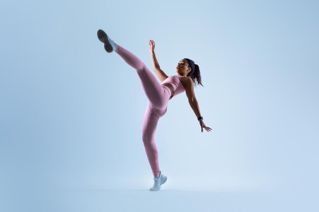 Fit black lady raising leg and showing perfect stretching doing karate side kick over blue background full length