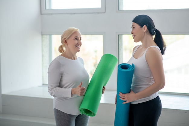 Fit blijven. Blonde vrouwelijke en donkerbruine vrouwelijke het houden van yogadekens en het bespreken van iets