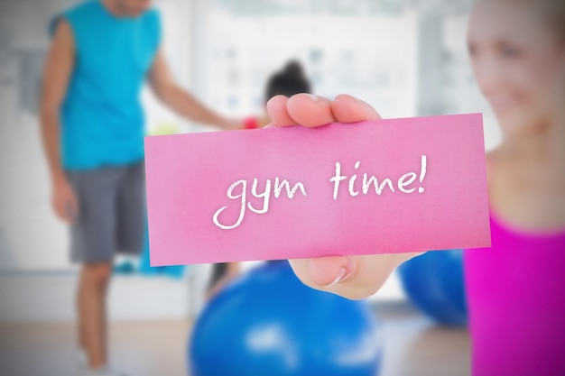 Fit blonde holding card saying gym time against trainer and client in fitness studio
