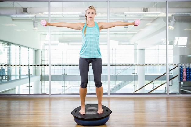 Fit blonde standing on bosu ball and lifting dumbbells in fitness studio