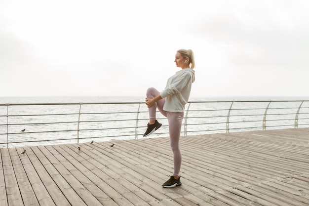 Fit blonde woman practicing leg stretching before outdoor workout. Young girl dressed in white hoodie trains on beach. Healthy lifestyle, sports and fitness