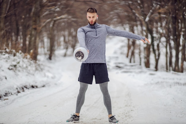 Fit bodybuilder standing on snowy path in forest and swinging kettlebell at winter. Bodybuilding exercises, winter fitness, outdoor fitness